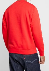 Röd sweatshirt med långa ärmar, rund halsringning och ribbade ärmslut. Mönsterfri tyg; bärs med mörka denimjeans som har bakfickor.