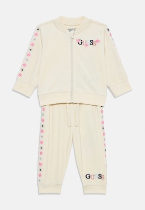 Cremefarvet velour tracksuit med lynlåsjakke. Har blomsterbroderi i pink og blå på ærmerne og bukserne. "GUESS" logoen vises.