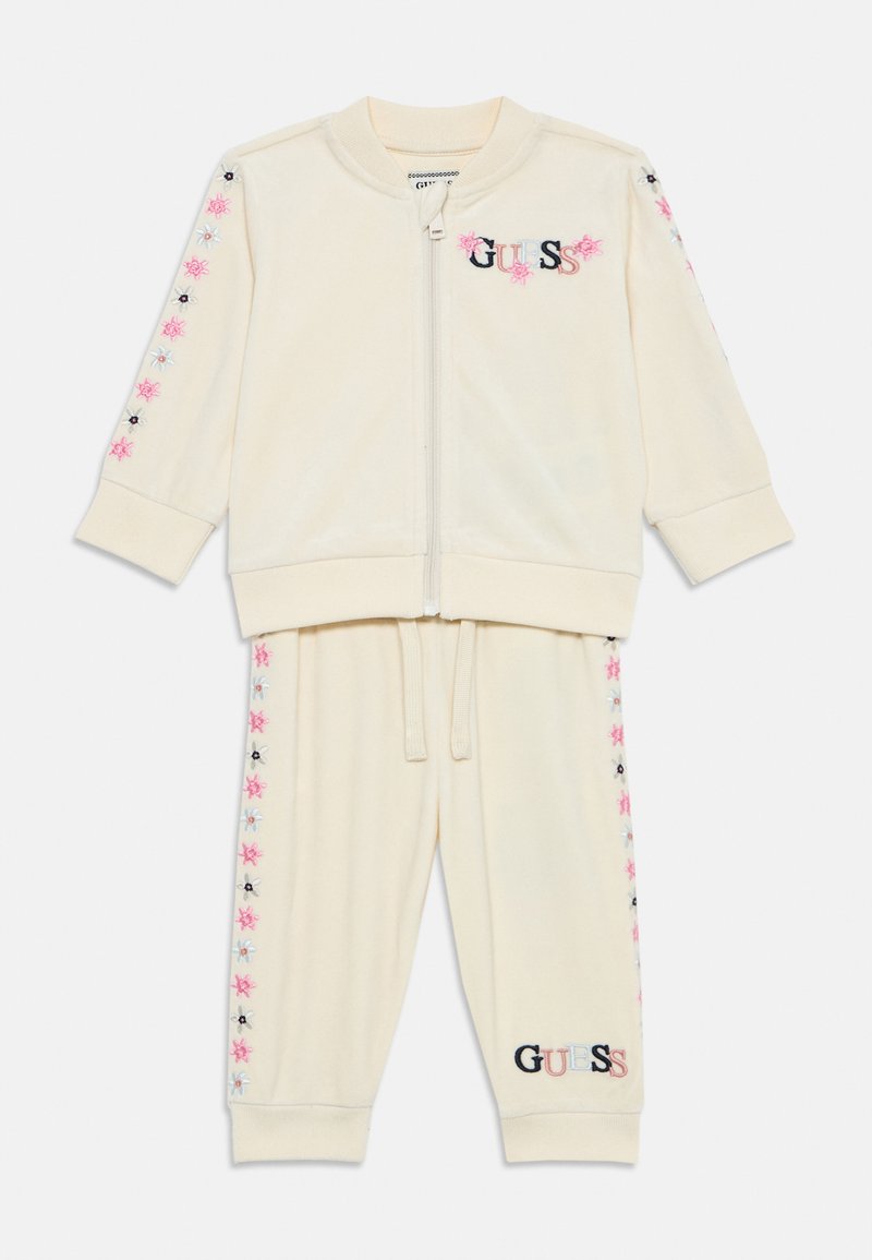 Tuta in velluto color crema con giacca con zip. Presenta ricami floreali in rosa e blu sulle maniche e sui pantaloni. Logo "GUESS" in evidenza.