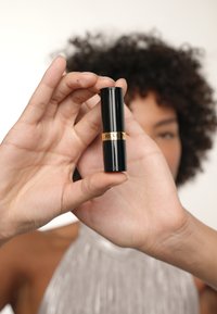 Rouge à lèvres Revlon dans un tube noir élégant avec des accents dorés, tenu entre des doigts. Le tube présente une finition lisse et brillante.