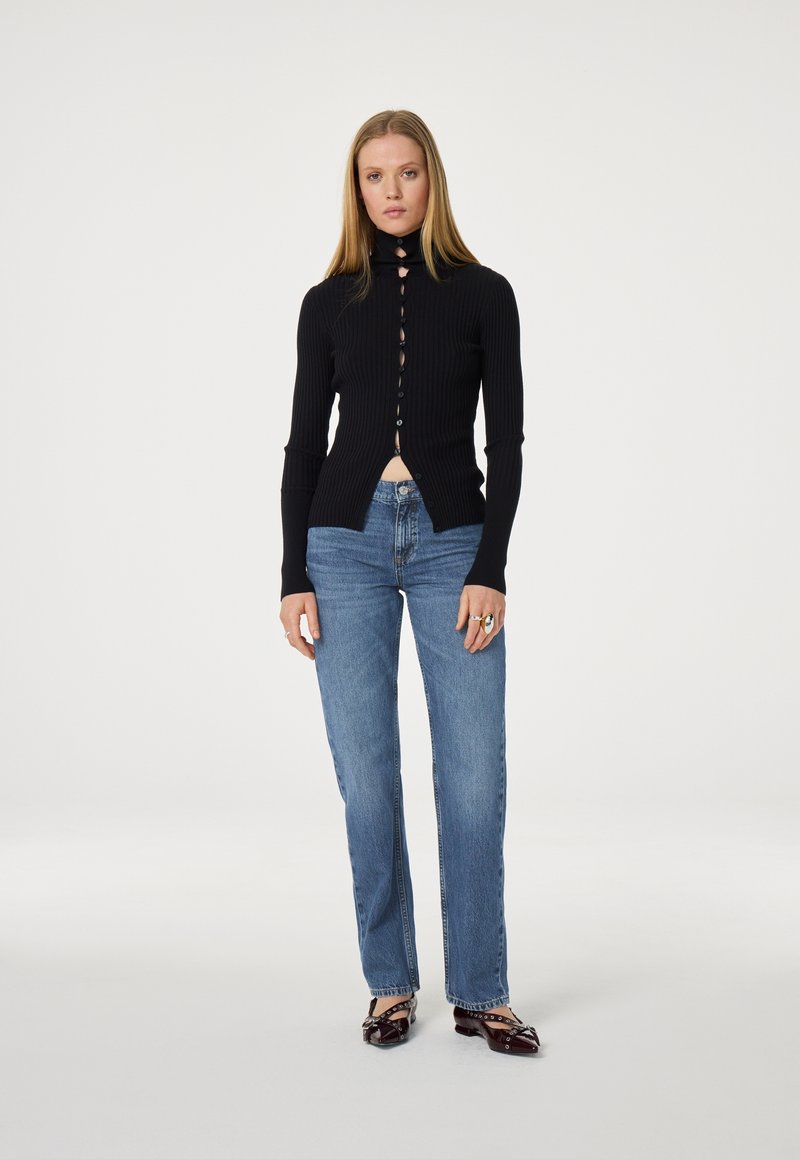 Zwarte geribbelde longsleeve top met hoge hals, knoopsluiting en uitsnijding, gecombineerd met blauwe recht gesneden jeans en bruine schoenen.