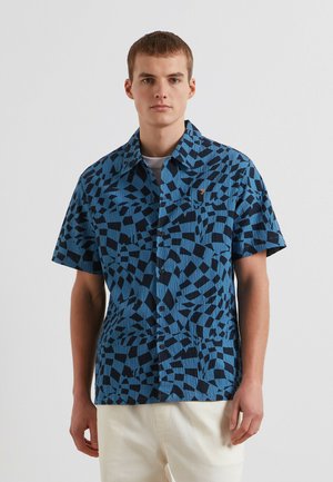 Giovane uomo con indosso una camicia a maniche corte con motivi geometrici blu e neri e pantaloni color avorio su uno sfondo semplice.