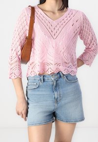 Rosa stickad topp med V-ringning, ärmar som slutar vid 3/4-längd och ett spetsigt, öppet mönster. I kombination med ljusblå denimshorts. En brun handväska syns.