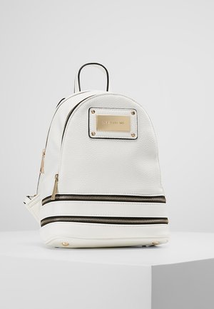 Malý bílý batoh s texturovaným materiálem, dvěma předními kapsami na zip, zlatými jezky zipů a zlatou značkovou destičkou na přední straně.