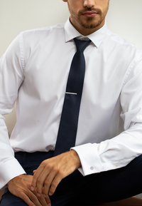 Camicia bianca a maniche lunghe con un motivo testurizzato, abbinata a una cravatta blu navy con un accento argento. Primo piano di mani su una superficie scura.