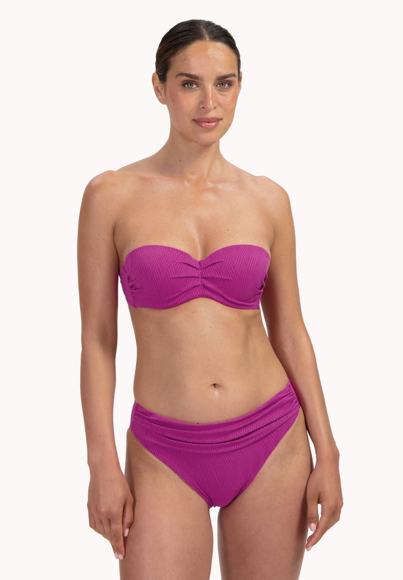 Kobieta w fioletowym bikini bez ramiączek z teksturowaną tkaniną, stojąca przodem na jasnym, jednolitym tle.