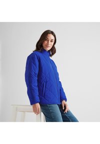 Woman wearing a bright blue quilted jacket and jeans, leaning on a white wooden stool against a plain light background.