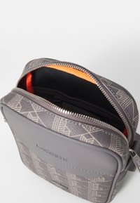 Gray Lacoste shoulder bag with a geometric pattern, zip closure, and orange lining. Features a smooth texture and adjustable strap.