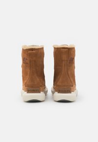 Bottines en daim marron avec un col en fourrure, semelles en caoutchouc et logo "SOREL" en relief au talon et sur le côté.