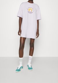 Ljuslila oversized t-shirtklänning med en stiliserad solnedgångsgrafik, kombinerad med vita strumpor med pastellrand och turkosa sneakers.