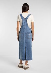 Džínová šaty s kšandami ve světle modré barvě, se šňůrkami na nastavování, dvěma předními kapsami a střední délkou s rozparkem vzadu pro pohodlný pohyb.