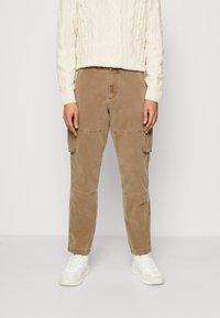 Persoon in beige cargobroek met flapzakken, witte sneakers en een crèmekleurige kabelgebreide trui, staande tegen een effen achtergrond.