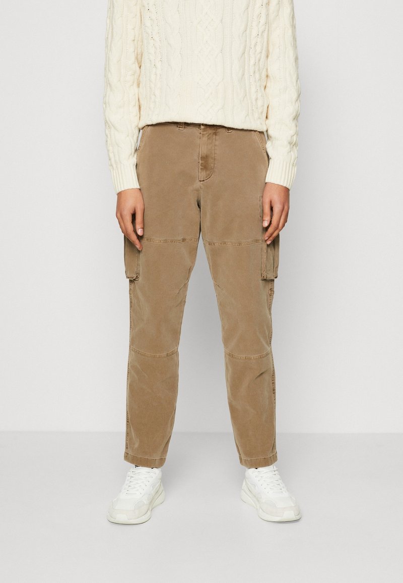 Persoon in beige cargobroek met flapzakken, witte sneakers en een crèmekleurige kabelgebreide trui, staande tegen een effen achtergrond.