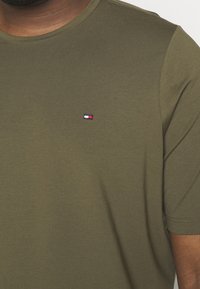 Muž v olivově zeleném tričku s krátkým rukávem, na levém prsu s malým obdélníkovým logem v červené, bílé a námořnické barvě.