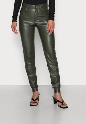 Modelka nosząca dopasowane ciemnozielone skórzane spodnie i czarne sandały na wysokim obcasie z paskami, stojąca na jasnoszarej podłodze na tle białej ściany.