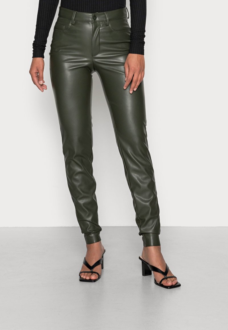 Mannequin portant un pantalon en cuir vert foncé ajusté et des sandales noires à talons hauts avec des brides, debout sur un sol gris clair contre un mur blanc.