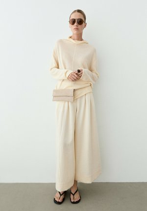 Frau trägt beigen lockeren Pullover und weite Hosen, Sonnenbrille, hält eine kleine beige Clutch, steht vor weißer Wand mit schwarzen Sandalen.