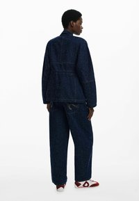 Giacca in denim blu scuro con vita aderente e maniche lunghe, abbinata a pantaloni a gamba larga. Entrambi presentano una trama maculata e cuciture a contrasto.