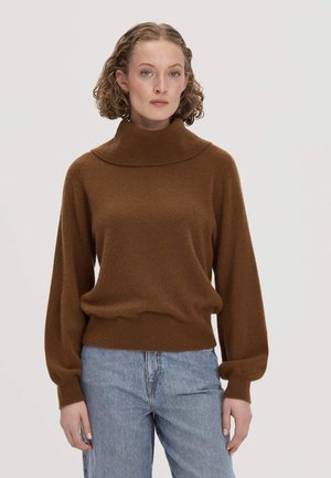 Femme aux cheveux bouclés portant un pull à col roulé marron et un jean bleu clair, debout devant un fond clair uni.