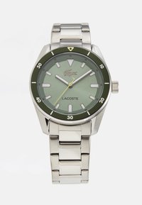 Ceas de mână din metal argintiu cu un cadran verde, markeri orari luminoși, brățară din oțel inoxidabil și o ramă neagră și verde. Dotat cu logo-ul Lacoste.
