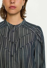 Camicia button-up grigia scura con strisce verticali bianche. Presenta un collo alto, maniche a palloncino texture e un design a zigzag sulla parte anteriore.