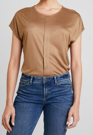 Kvinde iført en tætsiddende brun kortærmet top, der er stukket ind i højtaljede blå denimjeans med forlommer og knaplukning.