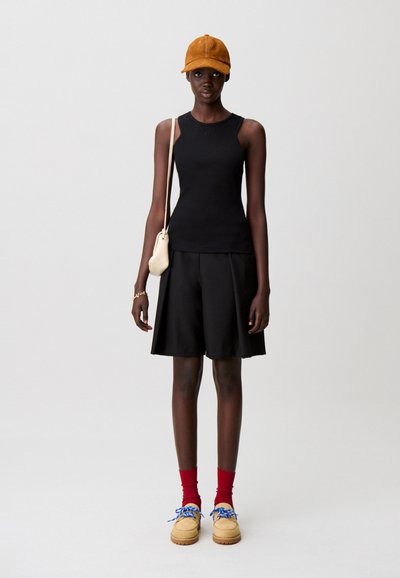 Top sin mangas negro, pantalones cortos plisados negros, gorra naranja, calcetines rojos y zapatos marrones con cordones azules. Pequeño bolso colgado sobre el hombro.