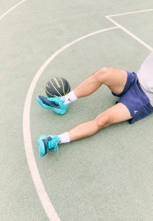 Osoba siedząca na zewnętrznym boisku do koszykówki, ubrana w turkusowe trampki i granatowe szorty, z czarno-żółtą piłką do koszykówki blisko stopy.
