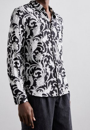 Mężczyzna ubrany w czarną koszulę z długim rękawem ozdobioną białymi kwiatowymi i winoroślami wzorami, w połączeniu z ciemnoszarymi spodniami.