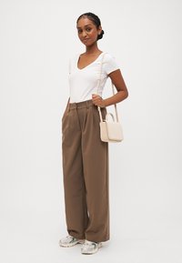 Modell som bär en vit V-ringad t-shirt och bruna högmidjade vida byxor, håller en liten beige handväska; grå sneakers med gröna accenter.