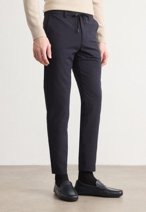 Mężczyzna noszący granatowe spodnie slim fit z regulacją w pasie na sznurek, czarne skarpetki i czarne mokasyny, stojący na neutralnej podłodze.