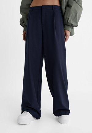 Osoba ubrana w szerokie granatowe spodnie i białe trampki, z widoczną częścią zielonej kurtki na jednolitym tle.