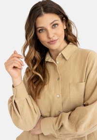 Beżowa koszula z kołnierzykiem, wykonana z fakturowanej tkaniny, z kieszonką na piersi i zapięciem na guziki, z lekko puffowanymi rękawami i luźnym krojem.