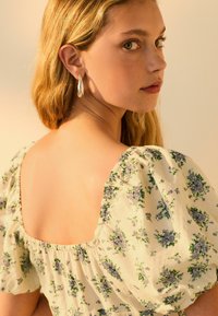 Gėlių rašto suknelė su pūstomis rankovėmis, turinti kvadratinę nugarą ir minkštą, struktūruotą audinį. Modelis dėvi sidabrinę ašarėlės formos auskarą.