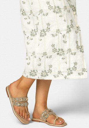 Füße, die beige verzierte flache Sandalen tragen, kombiniert mit einem weißen Rock mit grauer Blumenstickerei.