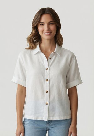 Mujer con cabello castaño ondulado, vistiendo una camisa blanca de manga corta abotonada y jeans azul claro, sonriendo sobre un fondo claro y liso.