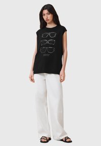 Schwarzes ärmelloses T-Shirt mit einem Grafikdesign von Sonnenbrillen in Weiß. Kombiniert mit lockeren weißen Hosen und schwarzen flachen Sandalen.