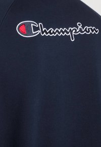 Námořnická modrá mikina s texturovanou tkaninou. Obsahuje vyšívané logo "Champion" v bílé a červené barvě na levé horní části hrudi.