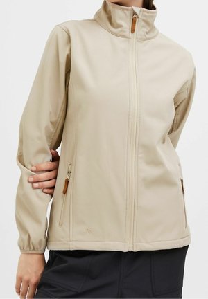 Beżowa kurtka na zamek z wysokim kołnierzem i zapinanymi na zamek kieszeniami, noszona na czarnych spodniach, model trzyma lewą rękę za plecami.