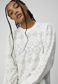 White lace blouse featuring geometric patterns, puffed sleeves, and a round neckline. The fabric is lightweight with a delicate texture.