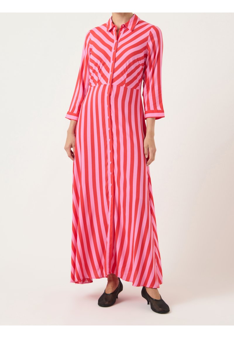 Femme portant une robe maxi à manches longues boutonnée avec des rayures diagonales et verticales rouges et roses, associée à des chaussures noires à talons bas.