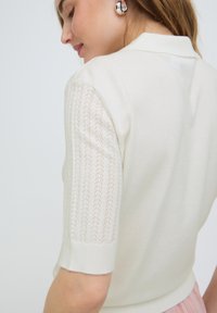 Femme en haut tricoté blanc à manches courtes texturées, boucles d'oreilles créoles argentées et jupe rose clair, de profil, les yeux fermés.