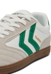 White and beige sneaker with green zigzag stripes, white laces, "hummel" logo in gold on side, and brown textured sole.