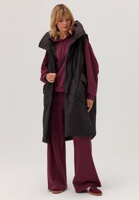 Kobieta w bordo bluzie z kapturem i szerokich spodniach, w dopasowanych sneakersach, nałożoną na długi czarny pikowany kamizelku, stojąca na tle jednolitym.