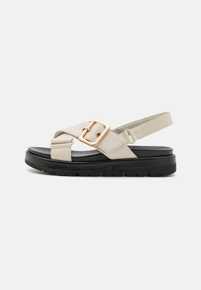 Beige leather sandal with cross straps and a gold hardware buckle. Black rubber sole with a thick, textured design. Adjustable slingback strap.