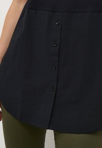 Camicia nera con bottoni, con orlo arrotondato e tessuto texturizzato, dotata di sei bottoni neri sulla parte anteriore. Indossata sopra pantaloni verdi con fantasia.