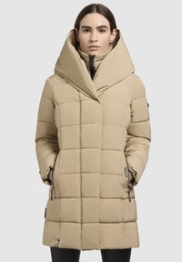 Kvinde iført en beige pivet dunjakke med stor hætte, hænderne i lynlåslommerne, der står imod en ensfarvet lys baggrund.