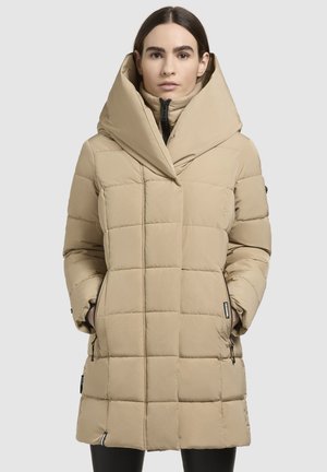 Egy nő bézs színű, steppelt pufidzsekiben áll, nagy kapucnival, a kezét a cipzáras zsebekben tartja, egy egyszínű világos háttér előtt.