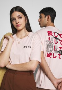Ljust rosa t-shirts med japansk text på framsidan och en ros-grafik med text på baksidan, tillverkade av mjukt bomullsmaterial.
