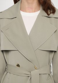 Trench-coat gris clair avec un design à double boutonnage et des boutons dorés, col large, et une ceinture assortie. Texture de tissu lisse.